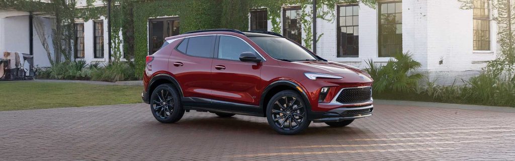 Front three-quarter view of a red 2025 Buick Encore GX parked on a brick driveway in front of a modern white building with greenery.