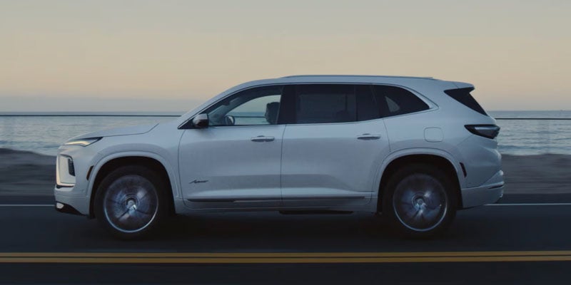 2026 Buick Enclave white luxury SUV driving along a coastal road with the ocean in the background.