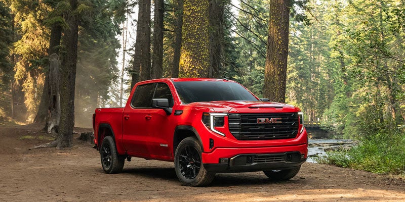 2026 GMC Sierra 1500 red pickup truck parked on a scenic forest trail surrounded by tall trees.