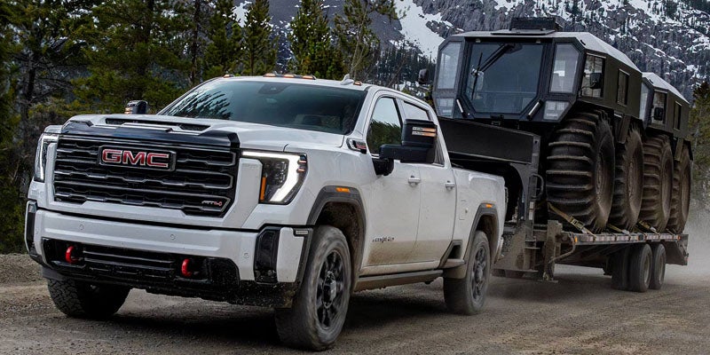 White GMC Sierra HD towing a large off-road vehicle on a mountain road.